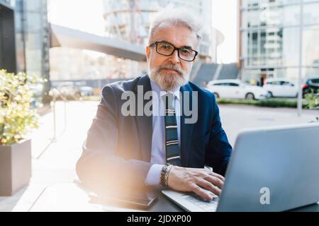 Senior businessman sitting with laptop outdoors Stock Photo