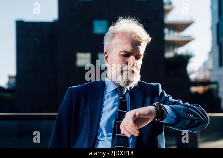 Senior businessman checking time on wrist watch standing outdoors Stock Photo