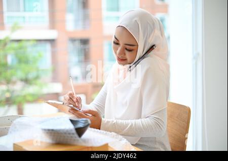 Professional and successful young muslim female small business owner talking on the phone with her client and noting an order on notepad. Stock Photo