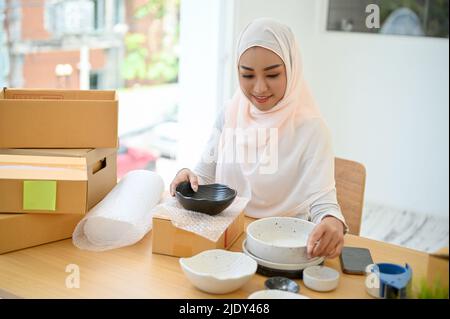 Successful asian muslim woman small business owner packing her kitchenware product into a shipping box to send to a customer. Stock Photo