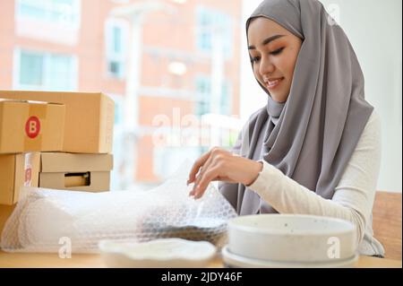 Attractive charming asian muslim female online e-commerce business owner preparing her customer's order, packing her product into a shipping box. Stock Photo