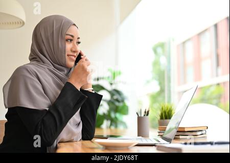 Gorgeous young asian muslim woman or businesswoman with hijab at the coffee shop, talking on the phone with her business partner. Stock Photo