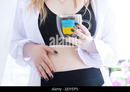 A pregnant girl holds a cup of coffee on which the flag of Ukraine is drawn, a new life during the war in the country, the victory of Ukraine, the fla Stock Photo