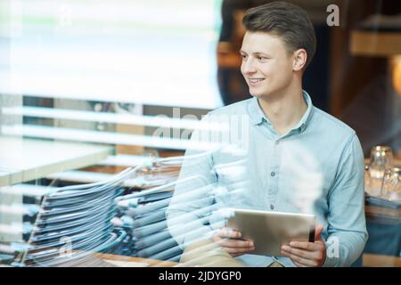 Smiling handsome young male social media specialist in casual shirt sitting in conference room and using tablet while presenting strategy at meeting Stock Photo