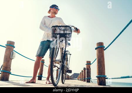 Portrait of a happy smiling barefoot man dressed in light summer clothes and sunglasses riding a bicycle on the wooden sea pier. Careless vacation in Stock Photo