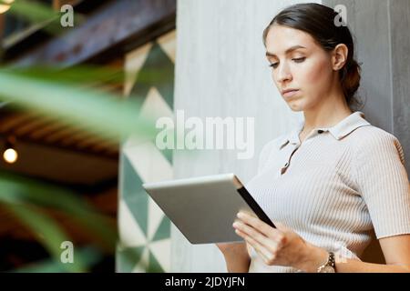Serious thoughtful young lady in beige shirt standing in coworking space hall and using tablet while searching for information Stock Photo