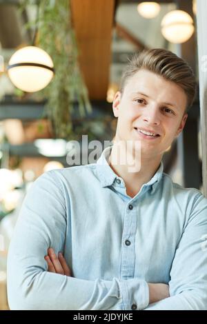 Portrait of smiling handsome young guy in light casual shirt leaning on wall and crossing arms on chest Stock Photo