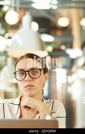 Portrait of serious confident attractive young lady in stylish spectacles using modern laptop in cafe Stock Photo