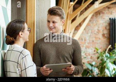 Smiling young handsome man in brown turtleneck using tablet while analyzing advertising ideas with colleague Stock Photo