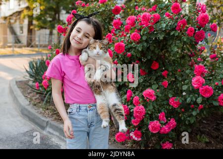 Cute kid girl 6-7 year old holding cat posing over blooming rose flower bushes in city street outdoor. Looking at camera. Childhood. Summer season. Fr Stock Photo