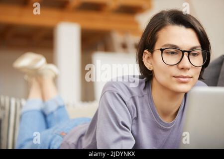 Content young female freelancer in eyeglasses lying on sofa and using laptop while working at home Stock Photo