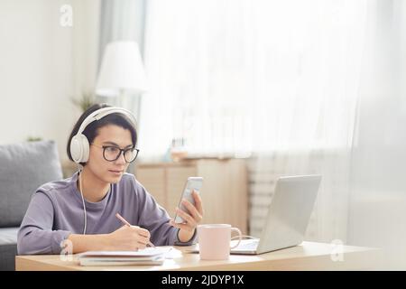 Serious curious attractive young woman in wired headphones sitting at coffee table and using smartphone while taking online course Stock Photo