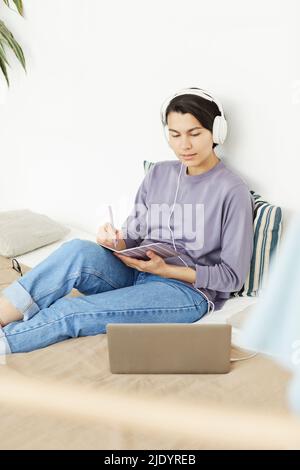Content curious young woman in headphones sitting on bed and making summary notes while listening to online course on laptop