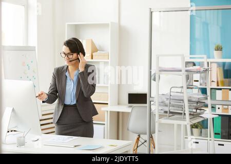 Content confident female manager in gray jacket standing at desk and discussing project while video conferencing with colleagues Stock Photo