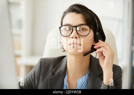 Portrait of serious confident beautiful female technical support operator in eyeglasses wearing headset with microphone for communication with clients Stock Photo