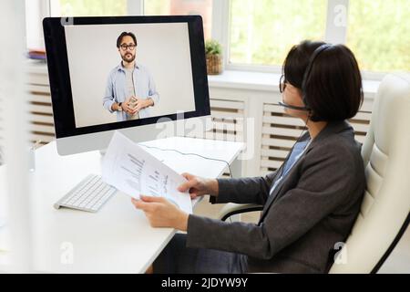 Serious young business analyst sitting at desk in office and analyzing statistics while interacting with Asian colleague via web conferencing provider Stock Photo