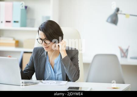 Displeased frowning female project manager in forma outfit sitting at desk with report paper and using laptop while talking via telecommunications app Stock Photo