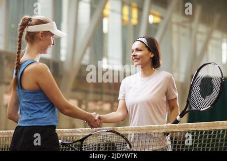 Smiling attractive mature woman in headband holding racquet and making handshake with tennis competitor at tennis court Stock Photo