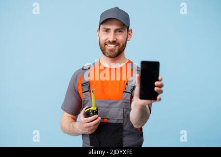 Smiling Man builder in uniform holding tape measure and showing empty screen phone in hands  Stock Photo