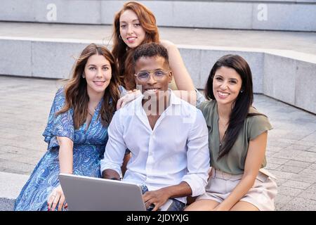 Group of multiracial people looking at camera and smiling while using a laptop outdoors on the street. Urban lifestyle and technology concept. Stock Photo