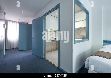 Cheap hotel room with blue carpet floors, serving hatch in the kitchen, and brown blankets on white duvets Stock Photo
