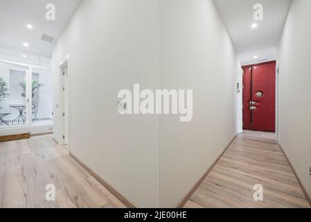 Distributor corridor with light chestnut flooring, entrance door upholstered in red skay and interior terrace with metal table and chairs Stock Photo