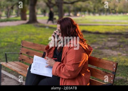An executive plus-size woman working outdoors. She is writing on her notebook and having a telephone conversation while sitting on a bench in a Stock Photo