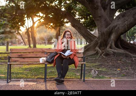 An executive plus-size woman working outdoors. She is holding a notebook and having a telephone conversation while laughing and sitting on a bench in Stock Photo