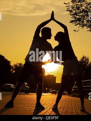 Young couple exercising on street near road in city from below view, sunset background. Darkened silhouettes of training man and woman looking to each Stock Photo