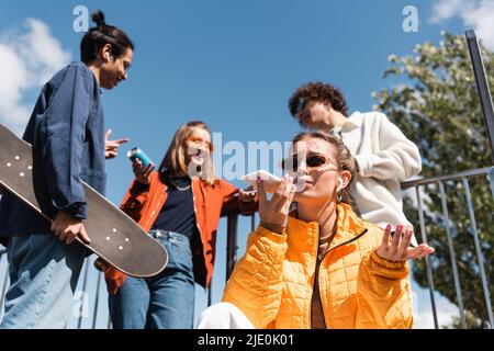 stylish woman sending voice message on smartphone near blurred interracial skaters Stock Photo