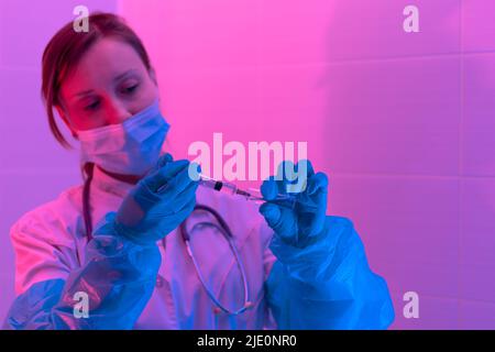 Beautiful young woman doctor with a stethoscope in a protective mask and rubber gloves in a bio laboratory with a syringe takes a sample of the virus. Stock Photo