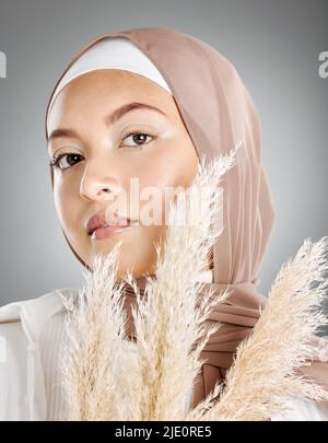 Studio portrait of one beautiful young muslim woman wearing brown headscarf posing with pampas wheat plant against grey background. Modest arab Stock Photo