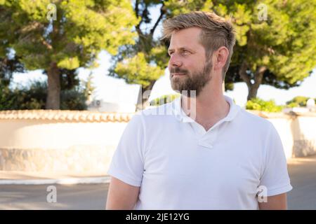 Outdoor shot of beautiful bearded young man posing over city garden on warm sunny day, wearing white polo shirt Stock Photo
