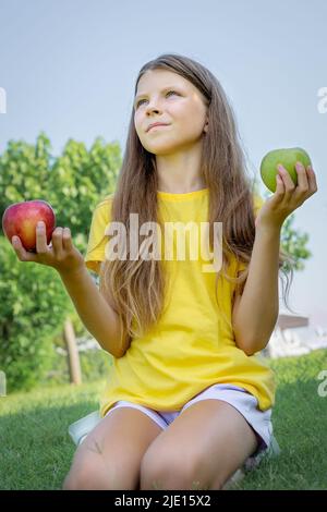A teenage girl holds apples in her hands while sitting on the green grass in the park. Stock Photo