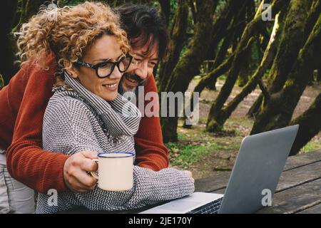 Man hugging woman while both using laptop in the outdoors nature park. Concept of couple in digital online travel leisure activity watching a computer Stock Photo