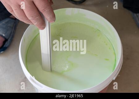 Worker's hands stir green paint before applying it, close-up. Bucket for mixing paints. Stock Photo