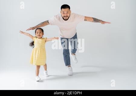 full length of cheerful asian father playing with daughter in yellow dress on grey Stock Photo