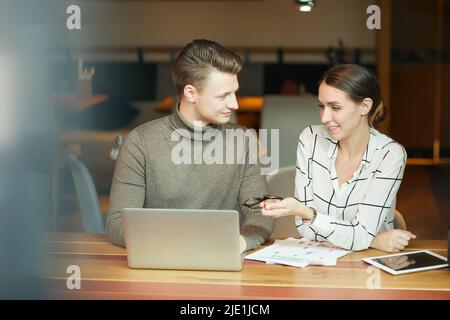 Positive young analysts sitting at table in coworking space and using laptop while working on business presentation together Stock Photo