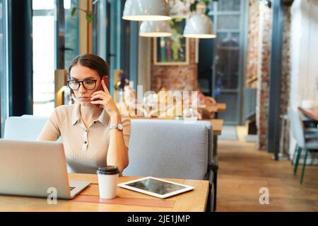 Serious multitasking young lady with fashionable wristwatch sitting at table in restaurant and discussing project with customer on phone Stock Photo
