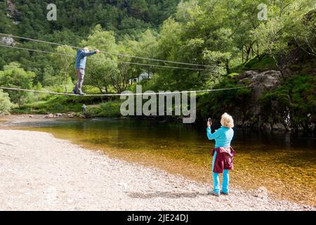 Woman photographing Man walking across Steall wire bridge over Water of Nevis river with Steall Falls Stock Photo