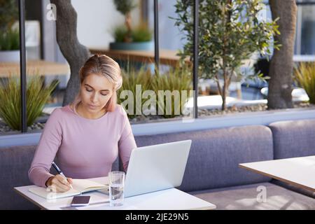 Concentrated young business lady sitting at table in modern cafe and making notes in organizer while planning meetings for day Stock Photo
