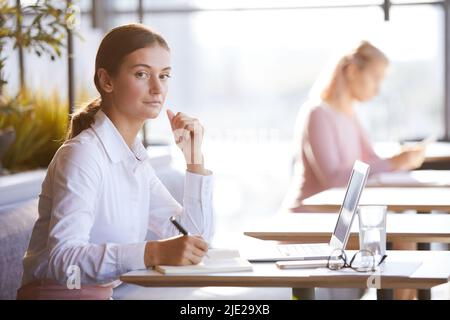 Portrait of serious attractive young businesswoman in formal blouse sitting at table in cafe and preparing for annual meeting Stock Photo