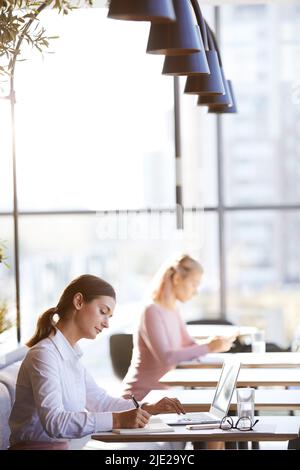 Serious beautiful young businesswoman in formal outfit sitting at table in cafe and making notes in diary while planning her day Stock Photo