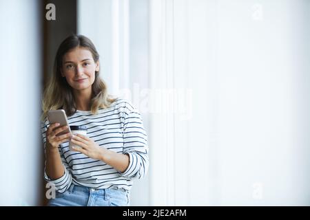 Portrait of smiling beautiful hipster designer in stripped sweater drinking takeout coffee and using smartphone Stock Photo