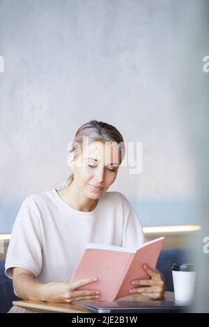 Content curious young lady sitting at table with coffee cup and spending time with interesting book Stock Photo