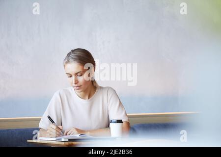 Serious concentrated blond lady in casual sweater sitting at table and making notes in diary while working in cafe Stock Photo