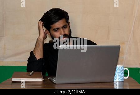 A young Indian man sitting on a chair and giving confused expression looking at laptop screen. Diary and coffee mug on table. Selective focus on face. Stock Photo