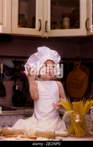 A kid in a cook's costume sits in the kitchen on a table and smiles Stock Photo