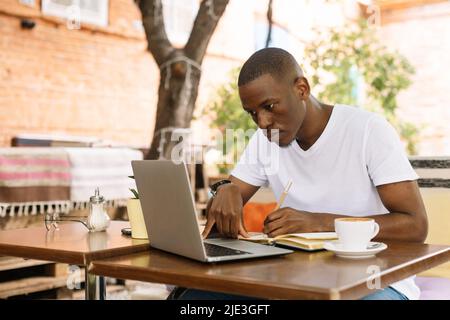 Serious, concentrated and enthusiastic multiracial man using laptop, writing notes in workbook in cafe. Student and exam Stock Photo