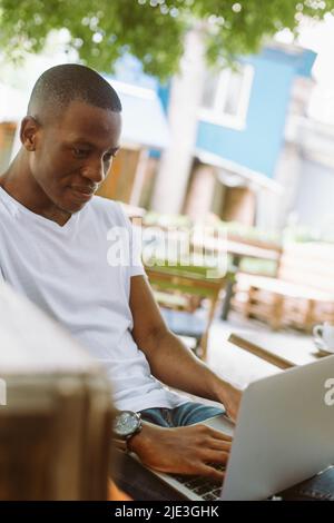 Concentrated multiracial man with laptop sitting on bench with legs in street coffee shop. Preparing to online webinar Stock Photo